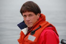 A person wearing a red and orange life jacket and a red jacket stands outdoors near a body of water, looking at the camera. The background is foggy or overcast.