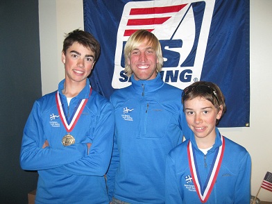 Three young people in blue jackets stand in front of a US Sailing flag. Two wear medals around their necks and all are smiling at the camera.