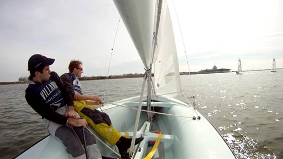 Two people are sailing a small boat on a sunny day, leaning out over the side to balance as they steer. Several other sailboats are visible in the background on the water.
