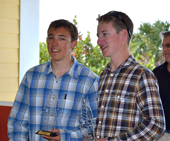 Two young men in plaid shirts stand side by side indoors, each holding a clear sailboat-shaped trophy. They are smiling, and an older man is partially visible in the background.