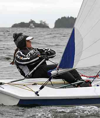 A person wearing a winter hat and jacket is sailing a small boat on a choppy body of water, holding onto the sail ropes, with tree-covered islands visible in the background.