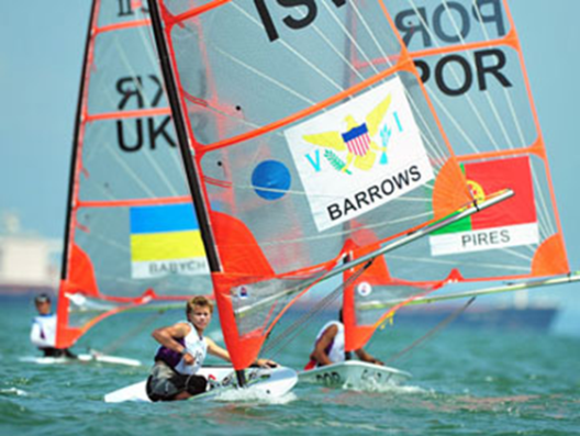 Three athletes compete in a sailing race, each on a small boat with large sails displaying country flags and names. The water is choppy, and ships are visible in the distant background.