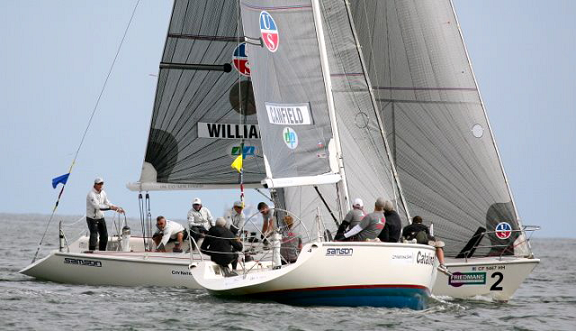 Two sailboats with crews compete closely on the water during a sailing race. The teams are actively managing the sails and steering as the boats lean into the wind. The scene is set on a calm, open body of water.
