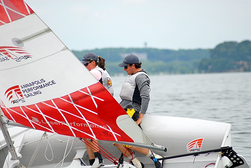 Two people in life vests and caps are sailing a small boat with a red and white sail on a calm body of water, with a forested shoreline visible in the background.