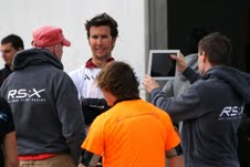 A man stands talking with three others, one of whom is filming or taking a photo with a tablet. Two people wear RS:X branded hoodies, and another wears a bright orange shirt. They are outdoors near a white wall.