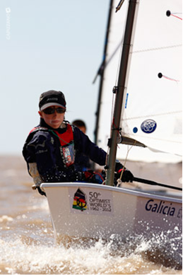 A person wearing sunglasses and a life jacket sails a small boat marked '50th Optimist Worlds 1962-2012' and 'Galicia' on the water, with sunlight sparkling on the waves.