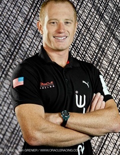 A smiling man in a black Oracle Racing polo shirt with a small US flag patch on the sleeve stands with his arms crossed in front of a textured, abstract background.