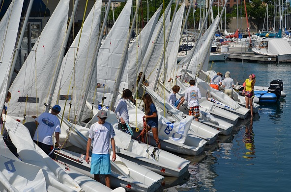 A group of people, mostly teenagers, prepare several small sailboats docked side by side at a marina on a sunny day, with more boats and water visible in the background.