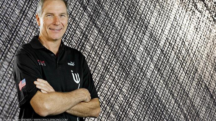 A man with gray hair wearing a black collared shirt with logos and an American flag patch stands with his arms crossed, smiling in front of a textured, crisscross-patterned background.