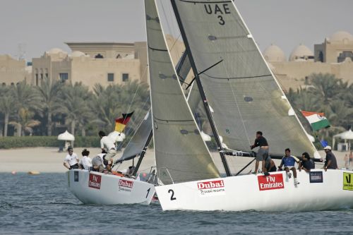 Two sailboats with crews racing on the water, displaying flags of Germany, UAE, and India; waterfront buildings and palm trees are visible in the background.