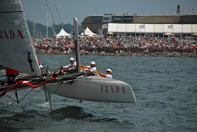 A sailing team races a Prada-branded catamaran on the water, leaning out over one hull as a large crowd watches from the shore on a sunny day.
