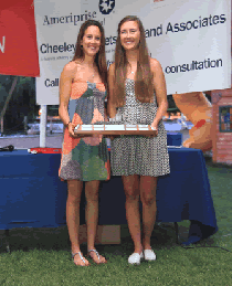 Two women stand side by side outdoors, smiling and holding a rectangular tray together. They are in front of a blue table and a banner with text, with grass and trees in the background.