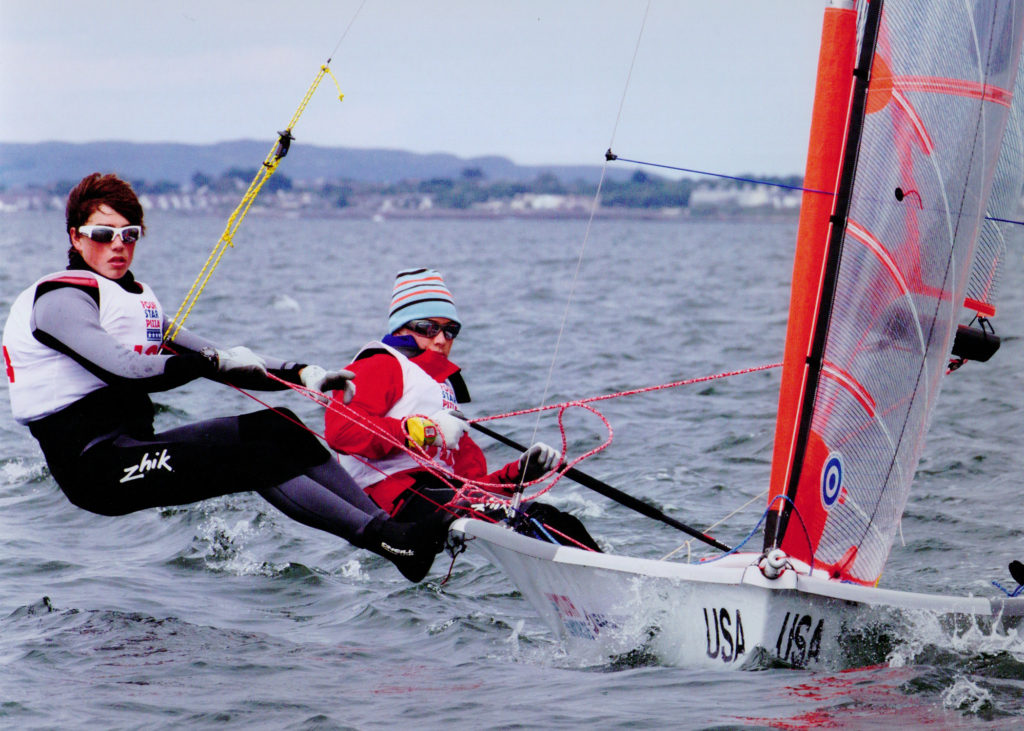 Two sailors in wetsuits and life jackets are racing a small sailboat marked 'USA' on the open water. One sailor leans far out over the edge, while the other steers. Distant shorelines are visible under a cloudy sky.