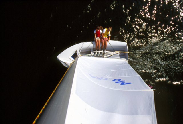 Aerial view of two people in life jackets sailing a small boat on dark water, with white sail billowing in the wind and sunlight reflecting on the water’s surface.