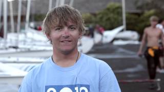 A young person with shaggy blonde hair wearing a light blue T-shirt stands outdoors by boats, with other people and sailing equipment visible in the background.