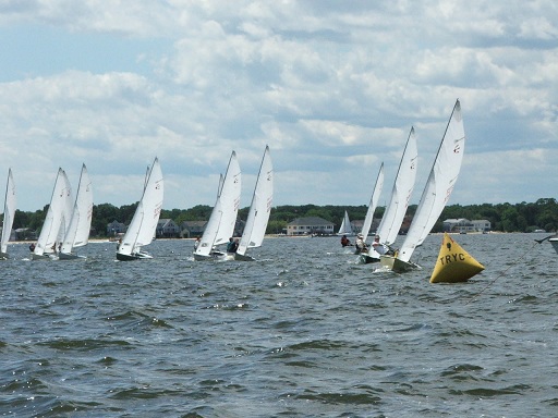 Several sailboats with white sails are racing on a lake near a yellow buoy marked 'JRYC,' with a shoreline and buildings visible in the background under a partly cloudy sky.