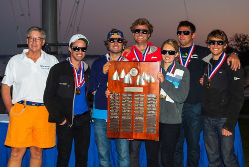 A group of six young adults wearing medals and sunglasses stand together, smiling, with one holding a large wooden trophy plaque. An older man in a white polo shirt and orange shorts stands beside them. It appears to be an outdoor event.