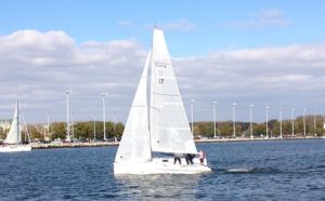 A white sailboat with three people aboard sails on a body of water near a shoreline lined with tall light poles and trees under a partly cloudy sky.