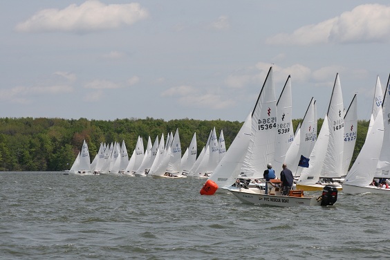A fleet of small sailboats with white sails lined up on a lake, preparing for a race. A motorboat with people aboard is in the foreground. Trees and a cloudy sky are visible in the background.