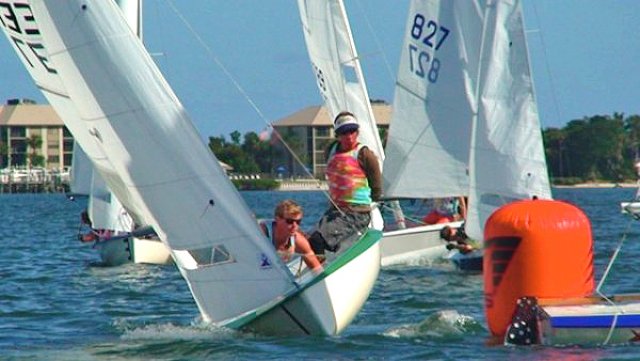 Two sailboats race near an orange buoy in a sunny coastal area. Sailors lean out to balance their boats, and buildings with palm trees are visible in the background.