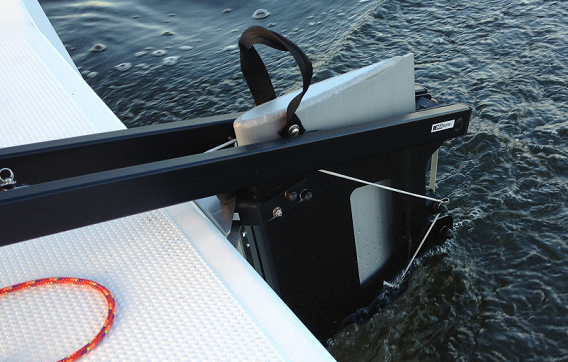 A close-up view of a boat's rudder partially submerged in water, attached to the stern with black metal fittings and a safety line, next to a textured white deck and an orange rope.
