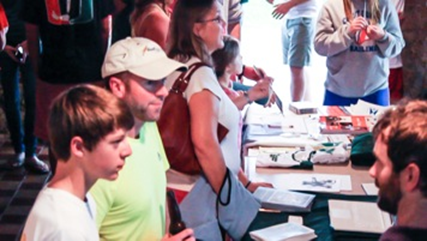 A group of people stand at a table covered with books and pamphlets, engaging in conversation at an indoor event. Some are holding bags and wearing casual clothing.