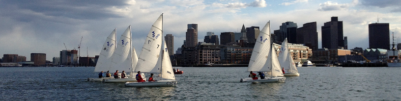 Several small sailboats with white sails are on the water, with people aboard, sailing near a city skyline of tall buildings under a cloudy sky.