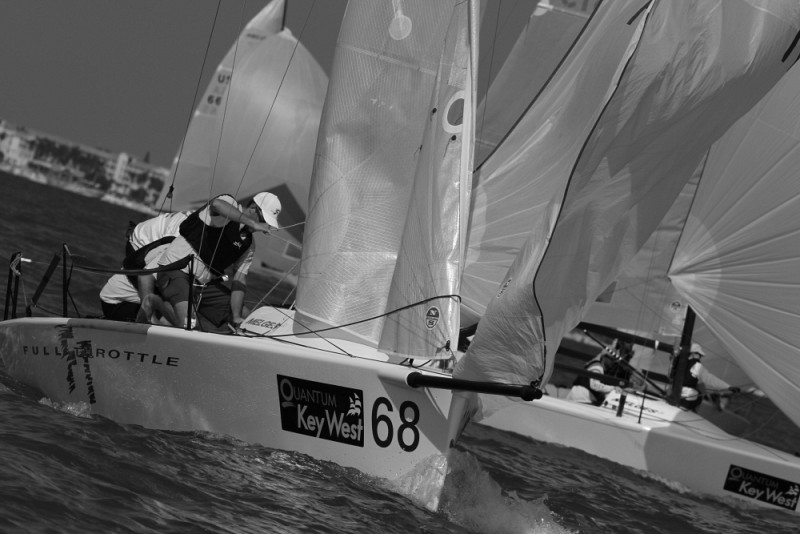 Black and white photo of two sailors maneuvering a sailboat numbered 68 during a regatta. The sails billow in the wind, with other sailboats and a shoreline visible in the background.
