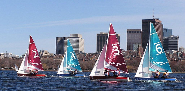 Four sailboats with numbered sails (2, 4, 3, and 6) race on a body of water with a city skyline and trees in the background under a clear blue sky.