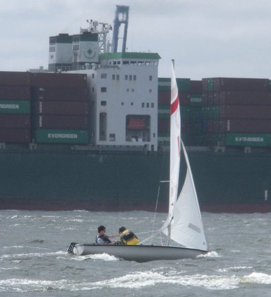Two people sail a small boat on choppy water while a large container ship, stacked with Evergreen shipping containers, passes in the background under a cloudy sky.