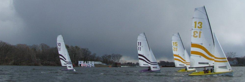 Four sailboats with large numbered sails (10, 11, 13, 15) race on a lake under a cloudy sky, with trees and buildings visible on the shore in the background.