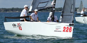Four people sail a white racing boat numbered 226 on calm water, wearing white shirts and caps. The boat displays Audi and Club logos, with another similar boat visible in the background.