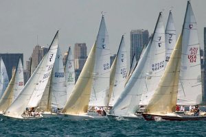 Several sailboats with white sails race close together on blue water, with a city skyline and tall buildings visible in the hazy background.
