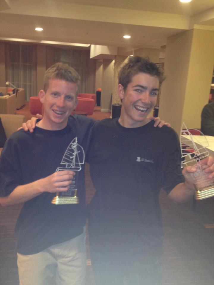 Two young men stand close together indoors, smiling and holding sailboat-shaped trophies. They appear happy and celebratory, with one having his arm around the other's shoulder.