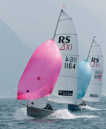 Two RS200 sailboats race across the water. The boat in front has a bright pink sail, while the one behind has a light blue sail. Both boats are cutting swiftly through the waves on a misty day.