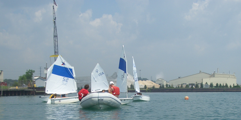 Three small sailboats with children wearing red life jackets are sailing near each other on calm water, with an industrial shoreline, buildings, and a crane visible in the background under a cloudy sky.