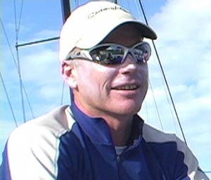 A man wearing a light-colored cap, reflective sunglasses, and a blue and beige jacket smiles outdoors with blue sky and sailboat masts in the background.