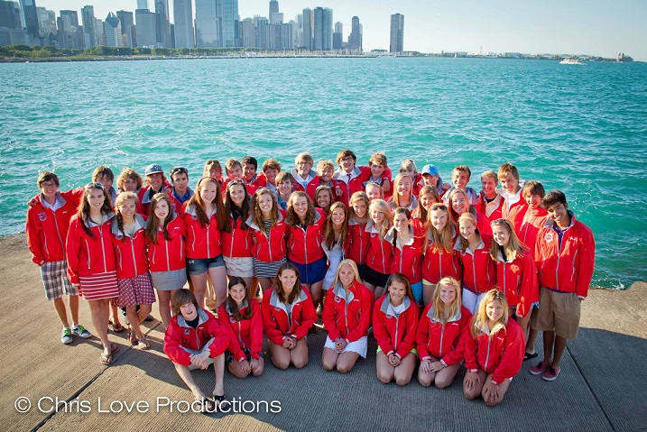 A group of about 40 young people in matching red jackets poses together on a lakeside promenade with a city skyline and tall buildings in the background under a clear blue sky.
