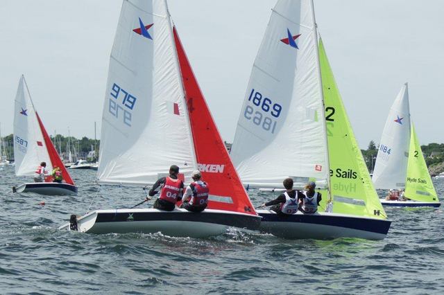 Several small sailboats with colorful sails and numbers compete on a lake. People in life jackets are sailing, and the background shows trees and a few distant boats under a cloudy sky.