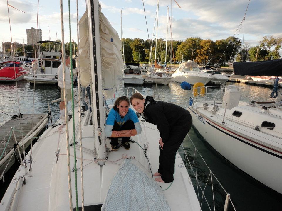 Two people are smiling and posing on the deck of a sailboat docked at a marina, with other boats, trees, and buildings visible in the background under a partly cloudy sky.