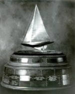 A black and white photo of a trophy featuring a model sailboat mounted on a large, round, multi-tiered base with engraved nameplates.