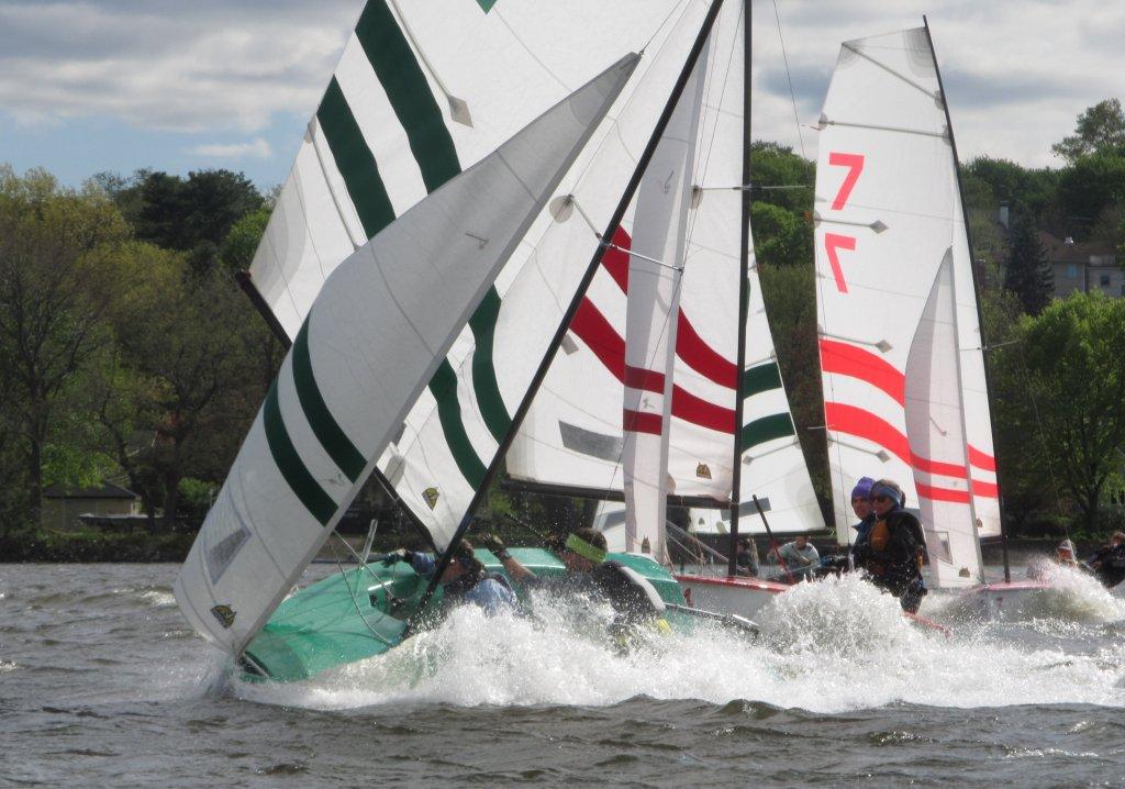 Several small sailboats with striped sails race closely together on a windy lake, causing splashes as they lean and cut through the water. Trees and houses are visible in the background under a partly cloudy sky.