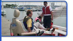 Two men wearing life jackets on a boat at a marina; one is seated and the other is standing, with sailboats and a dock visible in the background.