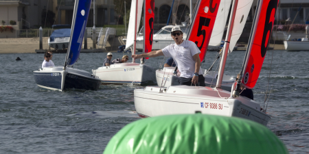 A man on a small sailboat points and shouts while competing with several other sailors on the water near a large green buoy. Other sailboats and buildings are visible in the background.