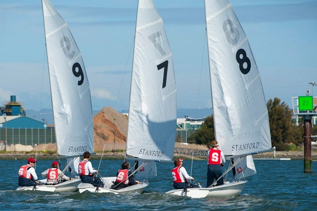 Three sailboats with white sails numbered 9, 7, and 8, each with a crew wearing red vests marked 'S,' race on a body of water near an industrial shoreline. The sails and boats are labeled 'Stanford.