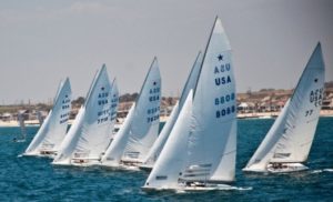 Six white sailboats with "USA" on their sails race closely together on blue water near a shoreline with houses and a sandy beach visible in the background under a partly cloudy sky.
