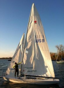 A person stands on a small sailboat with white sails on a calm body of water. The sails display the numbers “2859 6859” and a red star. Trees and another boat are visible in the background under a clear sky.