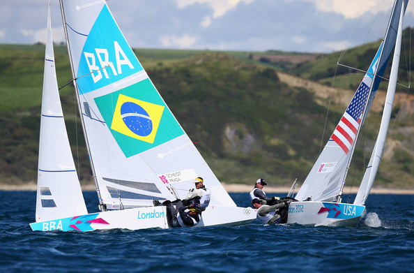 Two sailboats with Brazilian and American flags compete on the water at the London Olympics, with athletes onboard. A hilly, green shoreline is visible in the background under a partly cloudy sky.
