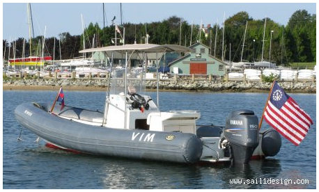 A grey inflatable motorboat named VIM with two outboard Yamaha engines floats on the water. It displays an American flag at the stern, with a marina and sailboats visible in the background.