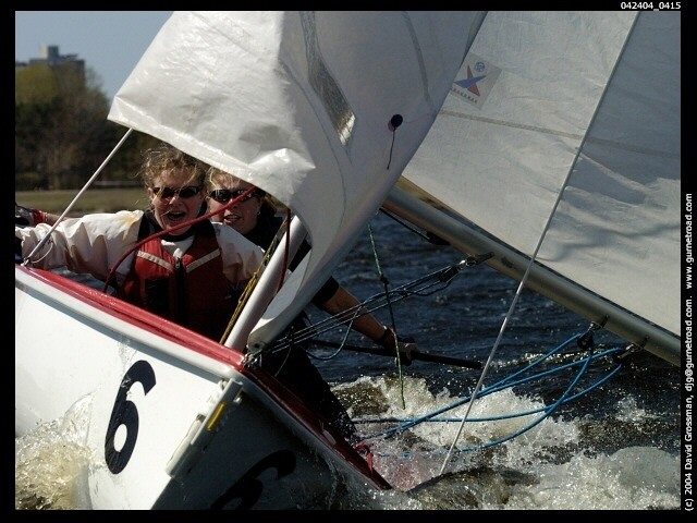Two people wearing sunglasses and life jackets are sailing a small boat numbered 6 on the water, leaning out as the boat tilts and splashes, with wind-filled sails and a shoreline in the background.